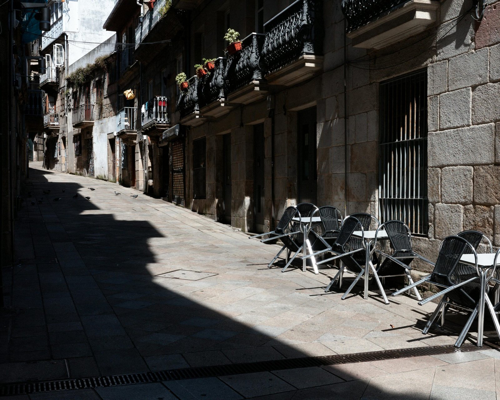 Empty street lined with overturned cafe tables, shadows, and flower-laden balconies.